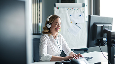 Woman dressed in business casual attire smiling while sitting at desk using computer with headphones on.