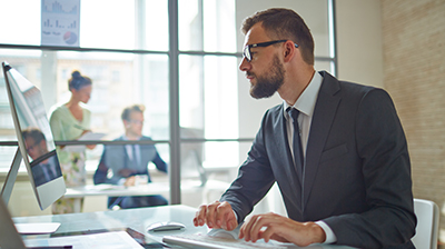 Man sitting in office using a computer with two people conversing in office next door.