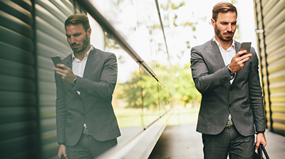 Man dressed in business attire walking outside while using phone and holding bag.