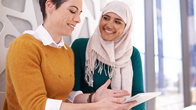 Two women smiling at each other while one woman is pointing at something on her tablet