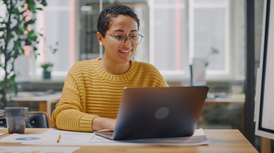 A woman in an orange sweater sitting at her desk and smiling while typing on her laptop.  