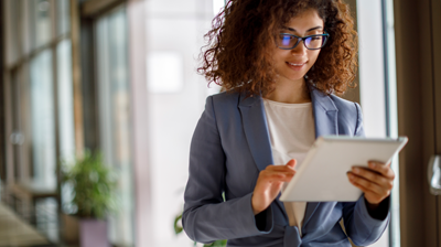 A woman dressed in business attire smiling while holding a tablet in one hand and scrolling on the tablet with her other hand.   