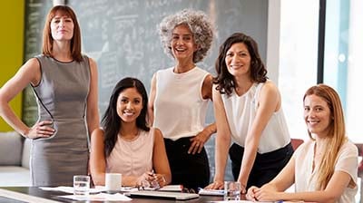 A group of five women dressed in business attire, standing and sitting around a table while smiling at the viewer 