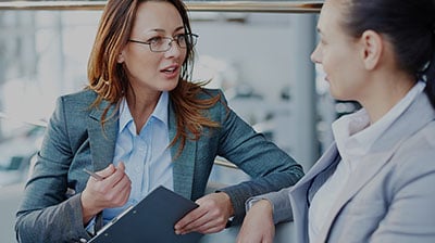 Two women in business attire facing each other and talking, one woman has a clipboard and pen in her hand.