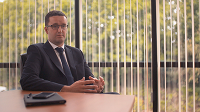 Man dressed in business attire sitting in conference room with large windows behind him.