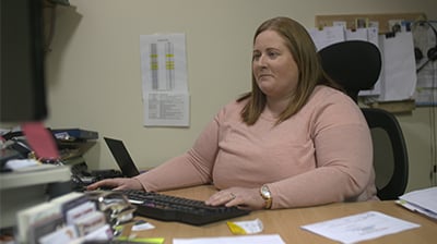 Woman in casual attire sitting behind a desk in an office, she is looking at a computer screen in front of her with her hand on a keyboard.