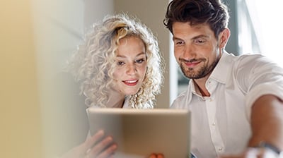 Woman and man dressed in casual attire smiling at tablet screen they are holding.