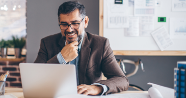 A man in business attire sitting at a table, he is smiling and looking down at an open laptop.