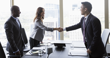 A group of people in business attire standing at a table in an office, a man and woman are shaking hands across the table.