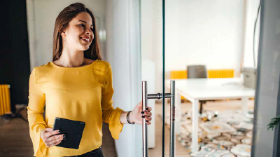 A woman in a yellow blouse, holding a tablet while opening a glass door 