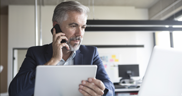 A man in business attire holding a cell phone to his ear and looking down at an open laptop in front of him.