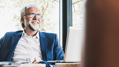 A man wearing a blue blazer smiling while sitting at a desk in front of a laptop 