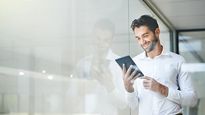 Man in business casual attire leaning on window and using tablet.