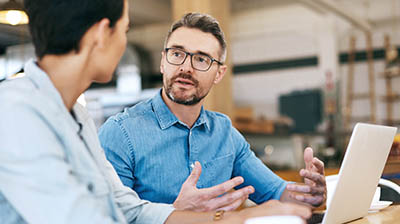 Two coworkers sitting at a desk and having a discussion while in front of a laptop  